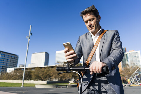 Älterer Geschäftsmann, der mit seinem Tretroller in der Stadt pendelt und sein Smartphone benutzt, lizenzfreies Stockfoto