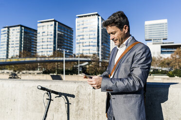 Mature businessman commuting in the city with his kick scooter, usine smartphone - GIOF05560