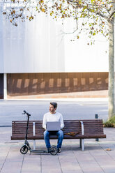 Lässiger Geschäftsmann mit Kick-Scooter, der auf einer Bank sitzt und entspannt in der Stadt arbeitet - GIOF05584