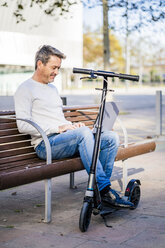 Casual businessman with kick scooter, sitting on a bench, working relaxed in the city - GIOF05589