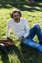 Lässiger Geschäftsmann, der eine Pause in einem Prak macht, im Gras sitzend, Musik hörend - GIOF05604