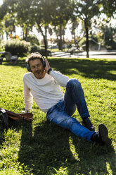 Casual businessman taking a break in a prak, sitting on grass, listening music - GIOF05605
