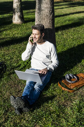 lässiger Geschäftsmann, der in einem Park im Gras sitzt, einen Laptop benutzt und telefoniert - GIOF05616