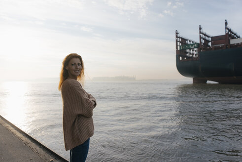 Deutschland, Hamburg, Frau steht auf Pier am Elbufer mit Containerschiff im Hintergrund - JOSF02891