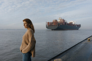 Deutschland, Hamburg, Frau steht auf Pier am Elbufer mit Containerschiff im Hintergrund - JOSF02893