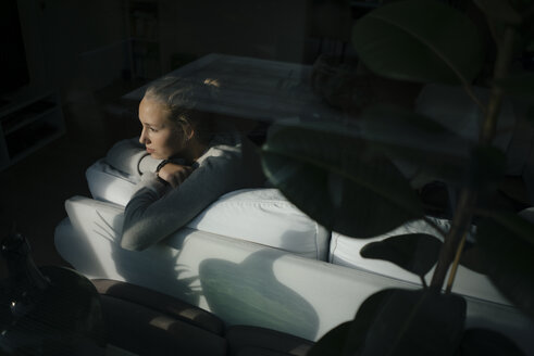 Serious teenage girl sitting on couch at home looking out of window - JOSF02940