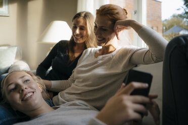 Happy mother with two teenage girls on couch at home with cell phone - JOSF02945