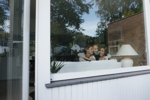 Glückliche Mutter mit zwei Mädchen im Teenageralter auf der Couch zu Hause hinter der Fensterscheibe, lizenzfreies Stockfoto