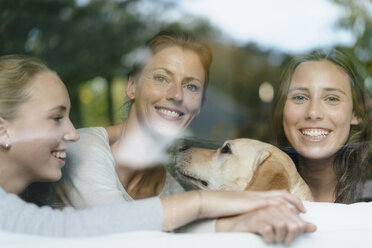 Porträt einer glücklichen Mutter mit zwei Mädchen im Teenageralter und einem Hund hinter einer Fensterscheibe - JOSF02954