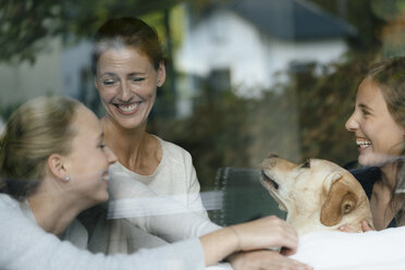 Glückliche Mutter mit zwei Mädchen im Teenageralter und Hund hinter einer Fensterscheibe - JOSF02955