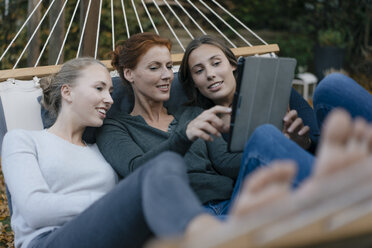Glückliche Mutter mit zwei Mädchen im Teenageralter liegt in der Hängematte im Garten im Herbst mit Tablet - JOSF02969