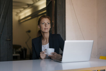 Nachdenkliche Geschäftsfrau sitzt am Schreibtisch im Büro mit einer Tasse Kaffee und einem Laptop - JOSF03006