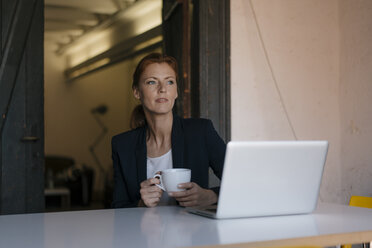 Thoughtful businesswoman sitting at desk in office with cup of coffee and laptop - JOSF03006