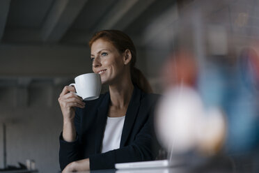 Lächelnde Geschäftsfrau mit einer Tasse Kaffee am Schreibtisch sitzend im Büro - JOSF03007
