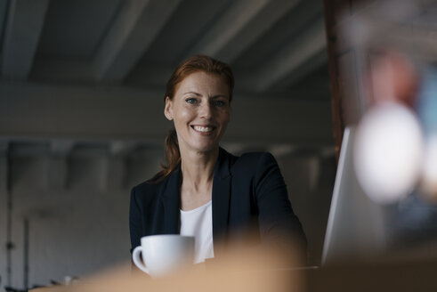 Porträt einer lächelnden Geschäftsfrau mit einer Tasse Kaffee, die am Schreibtisch im Büro sitzt - JOSF03008