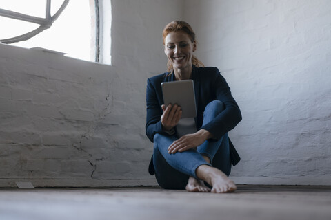 Barfüßige Geschäftsfrau, die im Büro auf dem Boden sitzt und ein Tablet benutzt, lizenzfreies Stockfoto