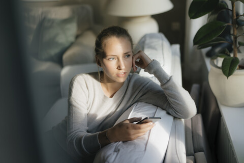 Serious teenage girl with cell phone sitting on couch at home stock photo