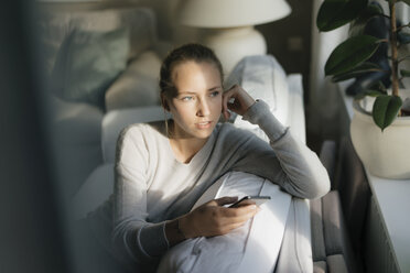 Seriöses Teenager-Mädchen mit Handy sitzt zu Hause auf der Couch - JOSF03050