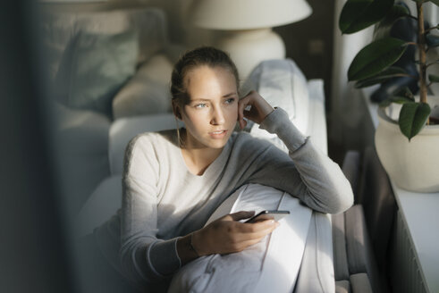 Serious teenage girl with cell phone sitting on couch at home - JOSF03050
