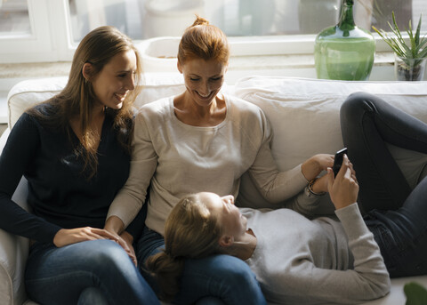 Glückliche Mutter mit zwei Mädchen im Teenageralter auf der Couch zu Hause mit Handy, lizenzfreies Stockfoto