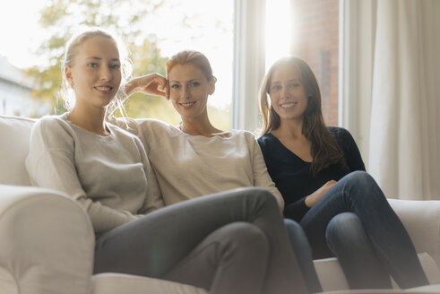 Porträt einer lächelnden Mutter mit zwei Mädchen im Teenageralter, die zu Hause auf der Couch sitzen - JOSF03053