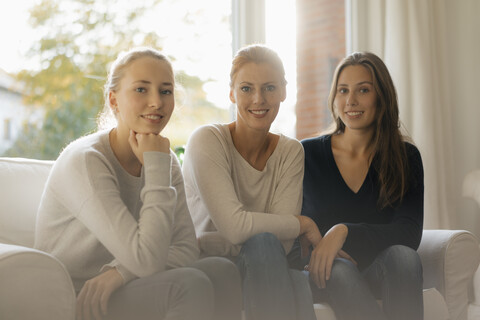 Porträt einer lächelnden Mutter mit zwei Mädchen im Teenageralter, die zu Hause auf der Couch sitzen, lizenzfreies Stockfoto