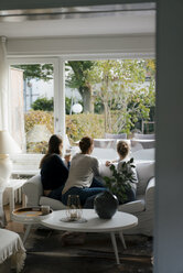 Rear view of mother with two teenage girls sitting on couch at home looking out of window - JOSF03058