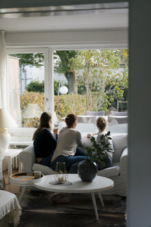 Rückansicht einer Mutter mit zwei Mädchen im Teenageralter, die zu Hause auf der Couch sitzen und aus dem Fenster schauen - JOSF03058