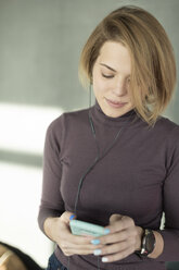 Junge Frau im Büro, die ein Smartphone mit Kopfhörern benutzt - VGF00208