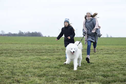 Zwei lachende Mädchen, die mit einem Hund auf einer Wiese rennen und einen Fung haben - ECPF00258