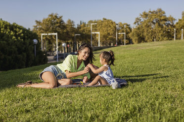 Glückliche Mutter spielt mit Tochter auf einer Decke im Park - MAUF02410