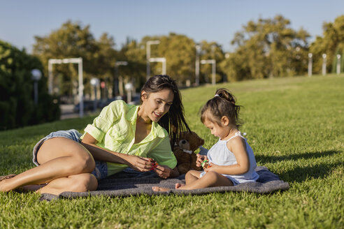 Happy mother playing with daughter on a blanket in park - MAUF02413