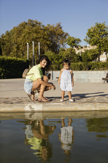 Happy mother with daughter looking at their mirror image in a pool in a park - MAUF02422