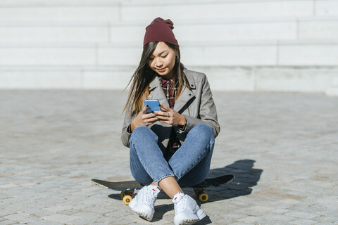 Lächelnde junge Frau sitzt auf ihrem Skateboard und schaut auf ihr Smartphone, lizenzfreies Stockfoto