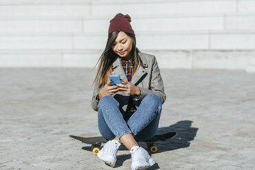 Smiling young woman sitting on her skateboard looking at smartphone - KIJF02207