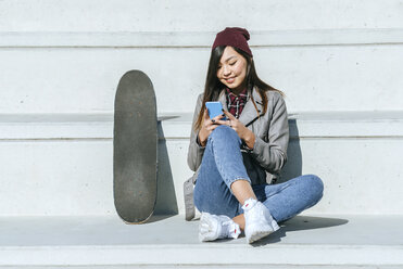 Lächelnde junge Frau sitzt neben ihrem Skateboard und schaut auf ihr Smartphone - KIJF02211