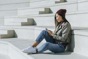 Lächelnde junge Frau sitzt auf einer Treppe und benutzt ein Smartphone - KIJF02212