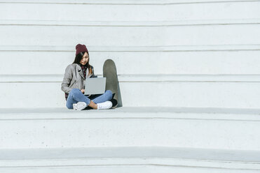 Junge Frau mit Skatboard sitzt auf einer Treppe im Freien und benutzt einen Laptop - KIJF02214