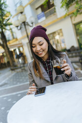 Porträt einer lächelnden jungen Frau, die in einem Straßencafé ihr Smartphone benutzt - KIJF02221