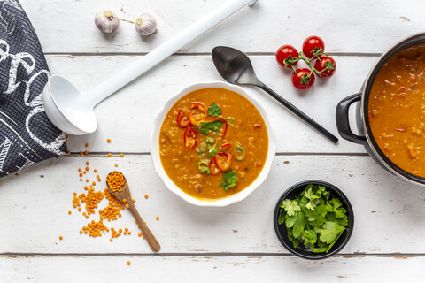 Schale und Topf mit garnierter roter Linsensuppe und Zutaten auf weißem Holz, lizenzfreies Stockfoto