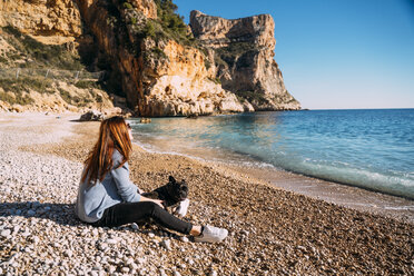 Spain, Alicante, Benitachell, redheaded woman sitting on the beach with her dog looking at view - OCMF00228