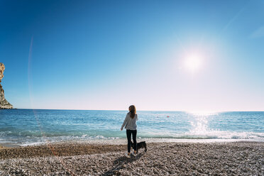 Spanien, Alicante, Benitachell, Rückenansicht einer Frau, die mit ihrem Hund am Strand steht und die Aussicht genießt - OCMF00229