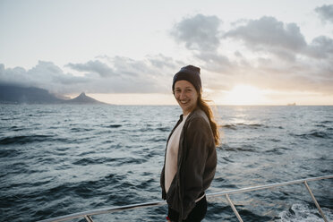 South Africa, young woman with woolly hat smiling during boat trip at sunset - LHPF00390