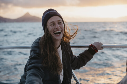 Südafrika, junge Frau mit Wollmütze lachend während Bootsfahrt bei Sonnenuntergang, lizenzfreies Stockfoto
