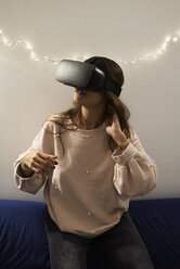 Young woman using VR headsets playing games on the sofa at home at night - IGGF00731