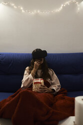 Junge Frau mit VR-Headset isst Popcorn auf dem Sofa zu Hause - IGGF00736