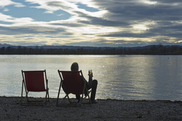 Deutschland, Mann sitzt am Ufer des Ammersees mit einem Glas Bier und beobachtet den Sonnenuntergang - CRF02823