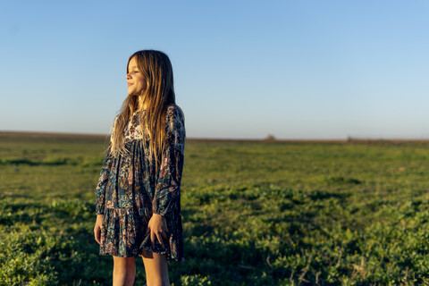 Lächelndes Mädchen im Kleid mit Blumenmuster steht in der Natur und genießt den Sonnenuntergang, lizenzfreies Stockfoto