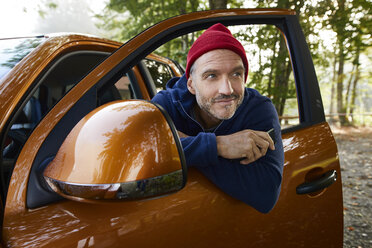 Portrait of bearded mature man wearing red cap leaning out of window of open car door - PNEF01185