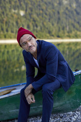 Portrait of bearded mature man wearing red cap and blue suit at lakeshore - PNEF01194